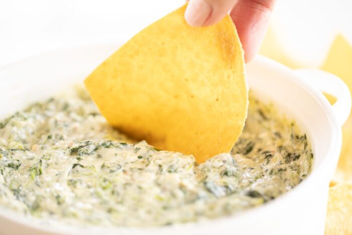 Fingers dipping a tortilla chip into a white bowl full of cream cheese spinach dip.