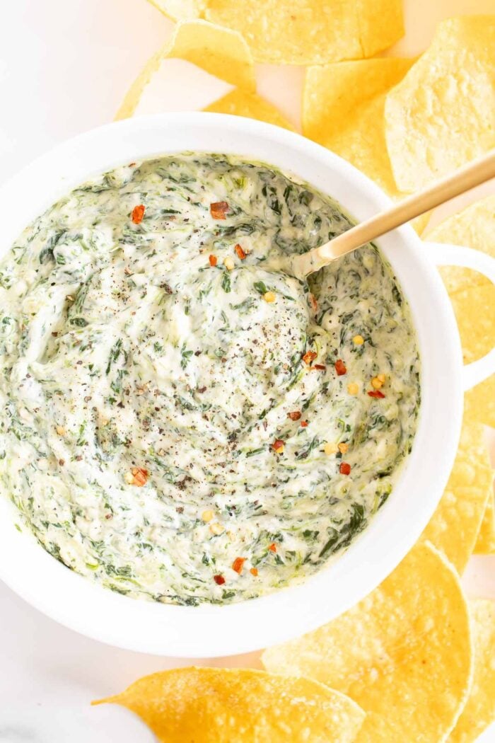 A white bowl full of spinach dip with cream cheese, gold spoon in the bowl and tortilla chips to the side.