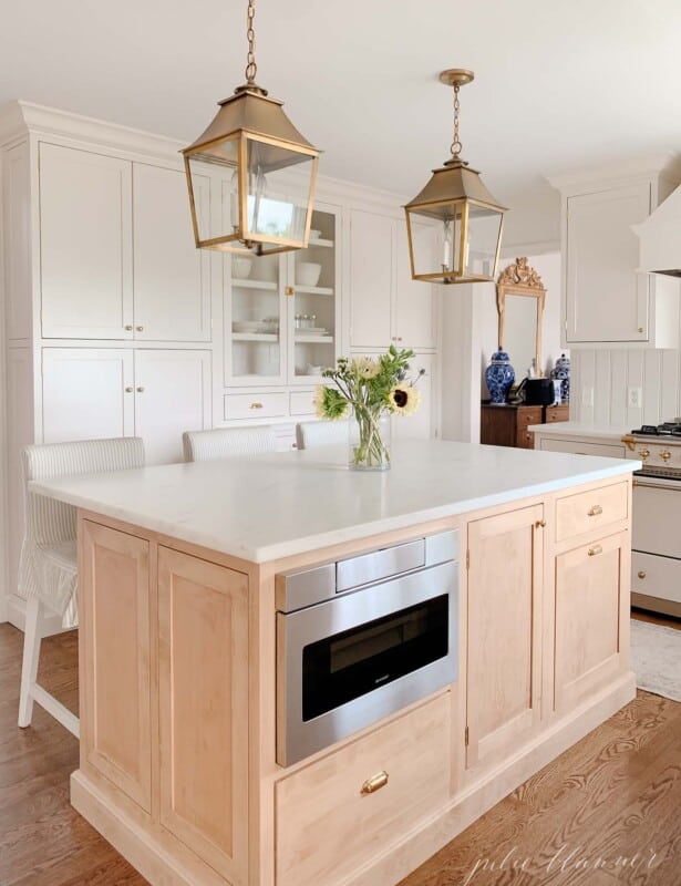 A soft wood toned kitchen island with a built in microwave drawer and brass lanterns above.