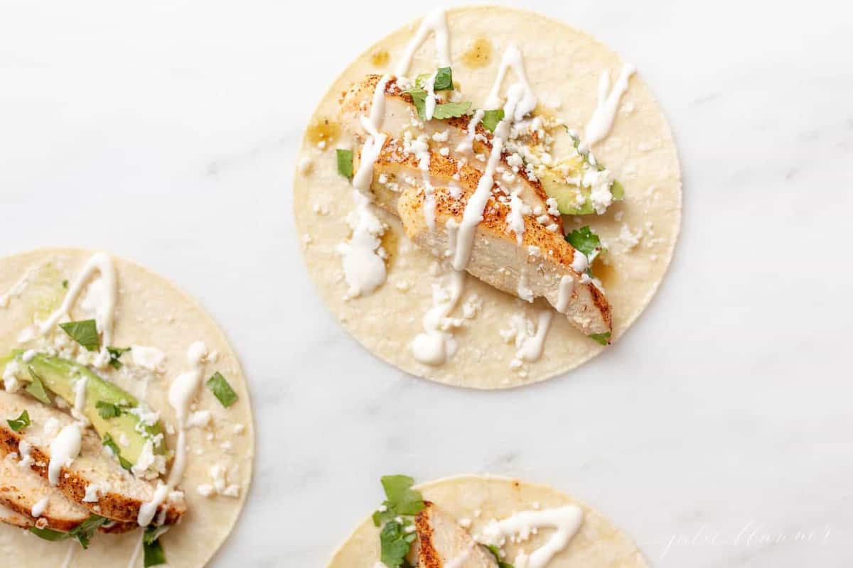White surface with three corn tortillas filled with chicken taco ingredients.