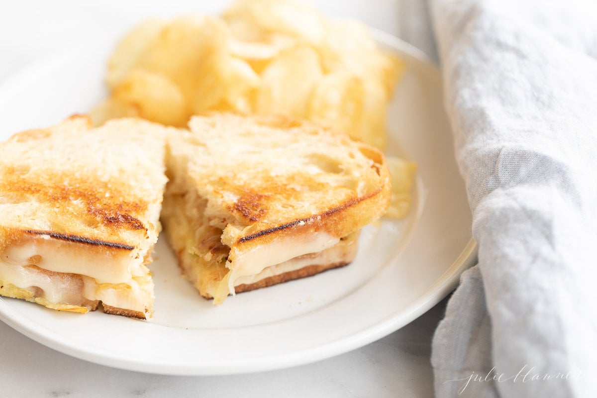 A white plate with a gourmet grilled cheese cut in half, chips in background.