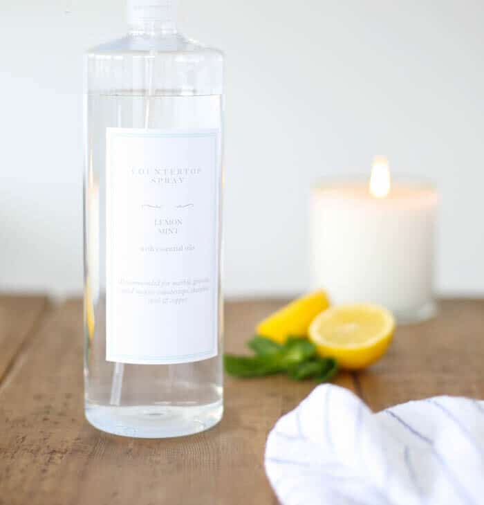 A clear glass spray bottle with a custom label, filled with natural all purpose organic cleaner - lemons and a candle in the background.