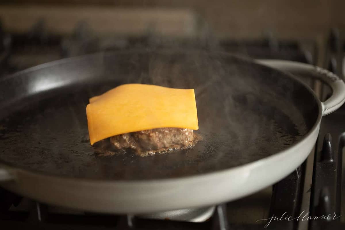 A burger cooking in a pan on the stovetop, with a piece of cheese on top. 