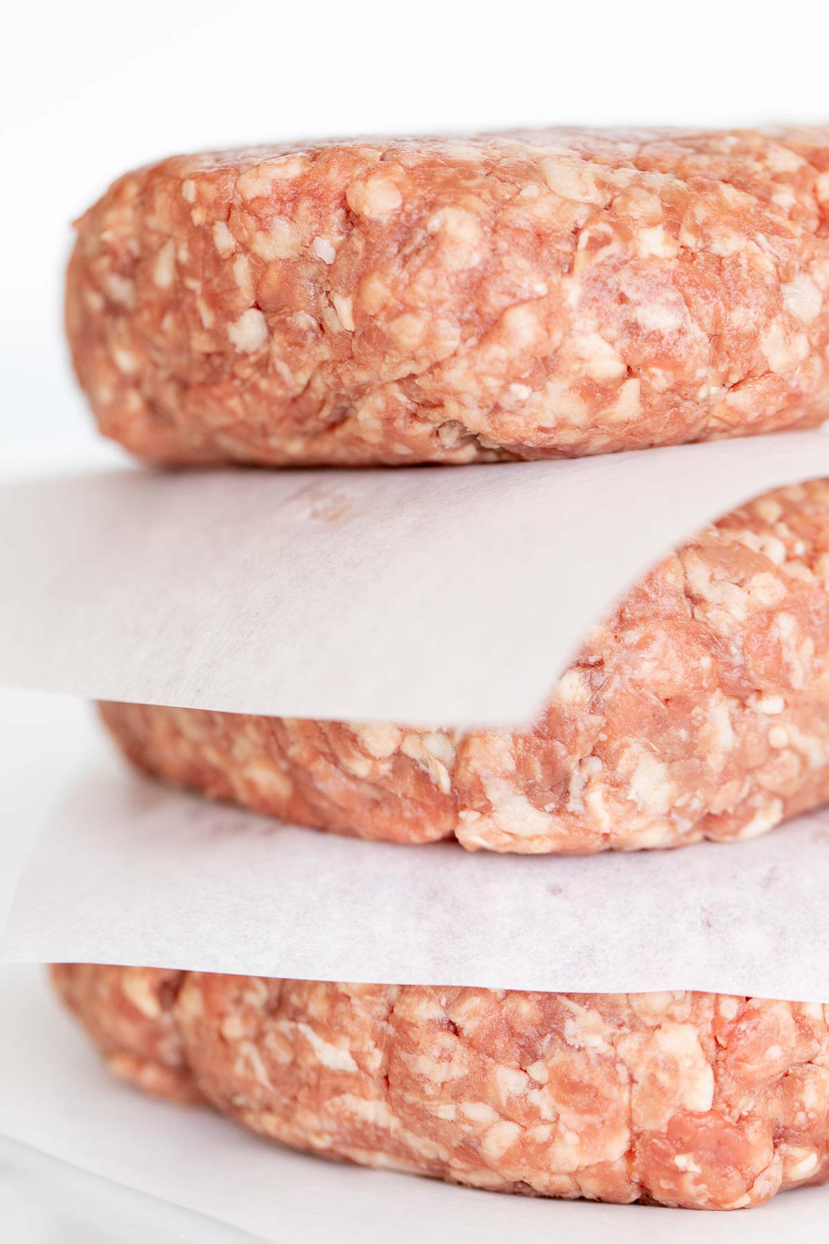 Close-up of three raw ground meat patties, ready to become juicy burgers, stacked with sheets of parchment paper between them on a white background.