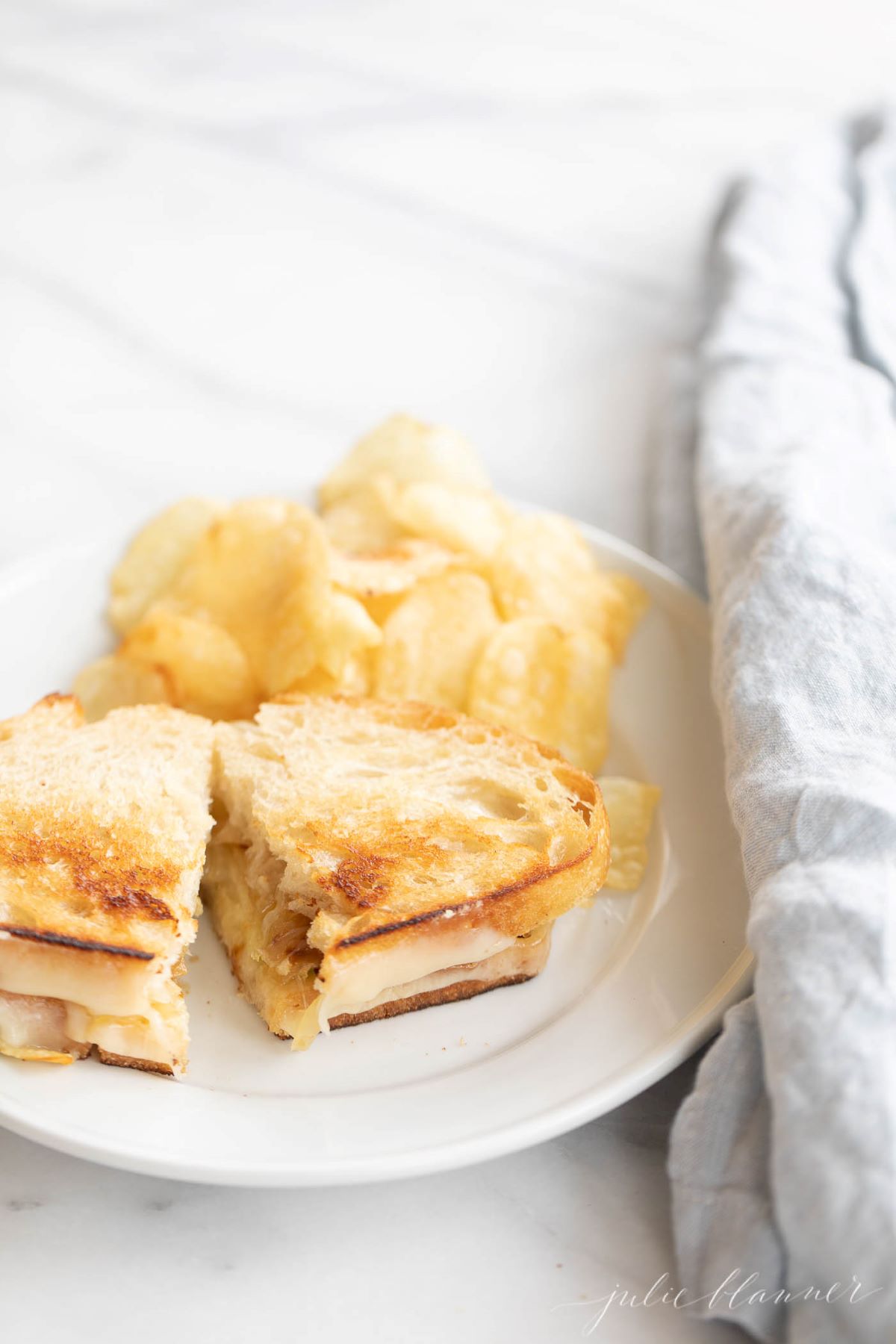 A white plate with a gourmet grilled cheese cut in half, chips in background.