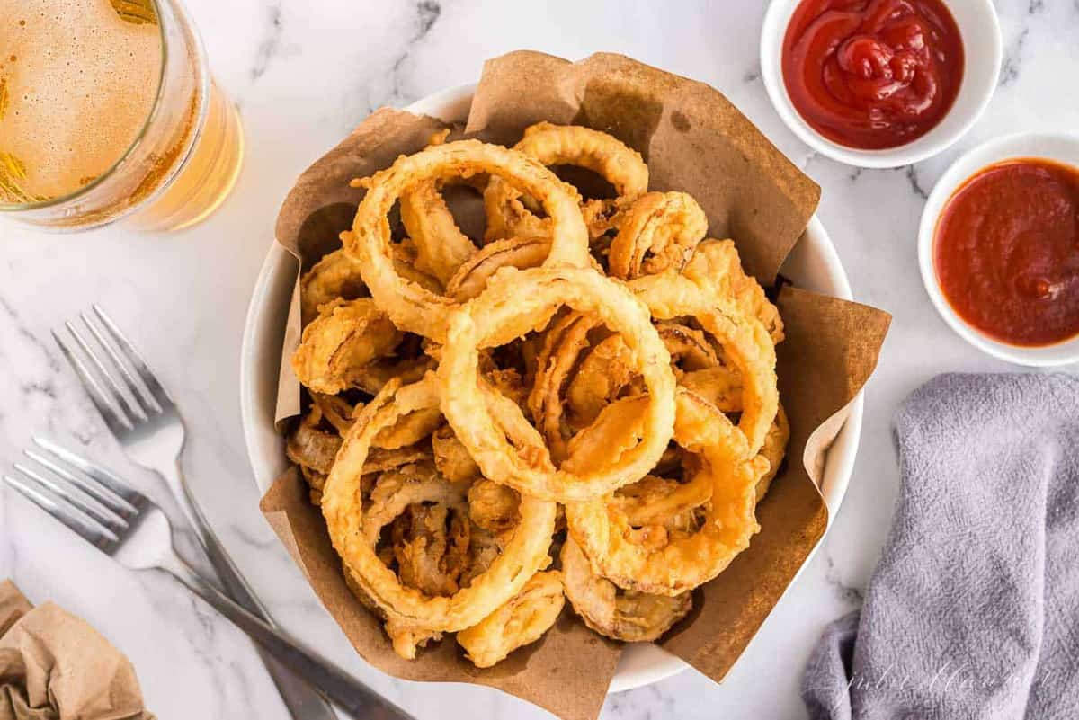 A white bowl of homemade onion rings, butcher paper lining the bowl, ketchup in the background.