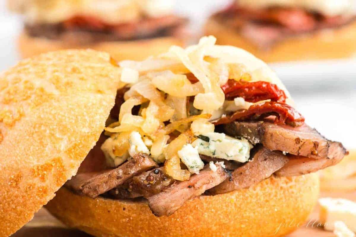 A steak sandwich with onions and blue cheese on top, bun balanced to the side.