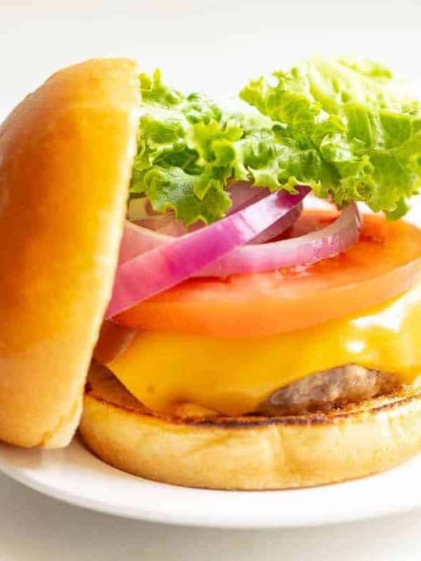 White background with a white plate featuring a classic cheeseburger, stacked with cheese, tomato, onion and lettuce, top bun to the side.