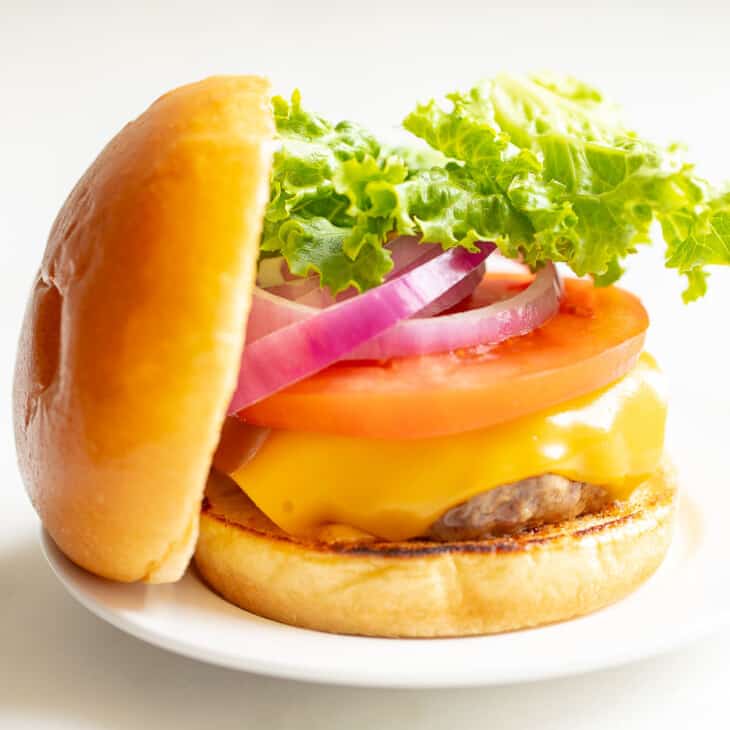 White background with a white plate featuring a classic cheeseburger, stacked with cheese, tomato, onion and lettuce, top bun to the side.