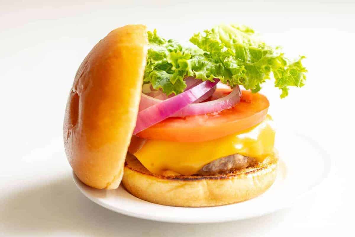 White background with a white plate featuring a classic cheeseburger, stacked with cheese, tomato, onion and lettuce, top bun to the side. 