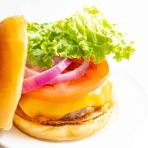 White background with a white plate featuring a classic cheeseburger, stacked with cheese, tomato, onion and lettuce, top bun to the side