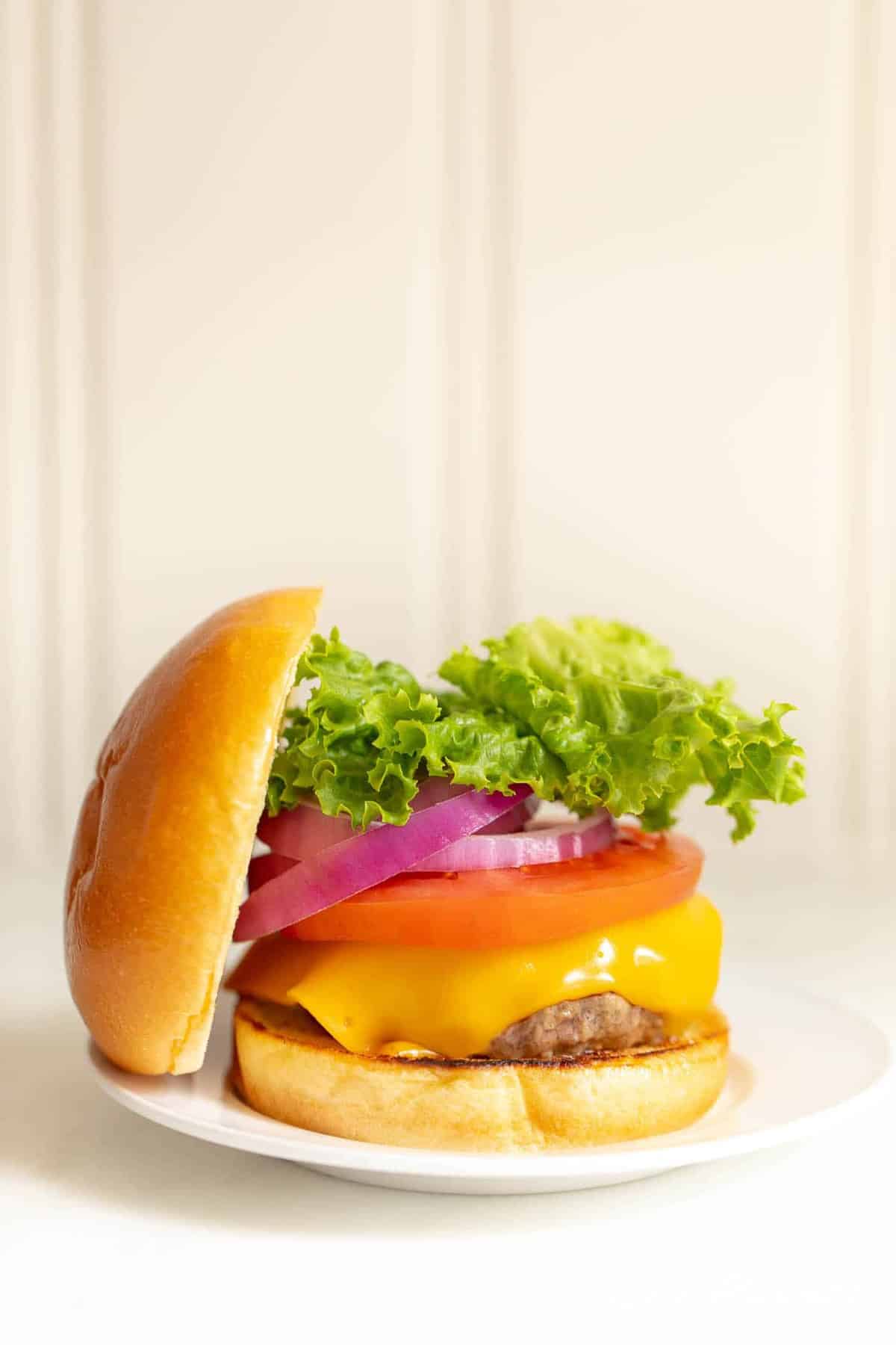 White background with a white plate featuring a classic cheeseburger, stacked with cheese, tomato, onion and lettuce with the top bun to the side. 
