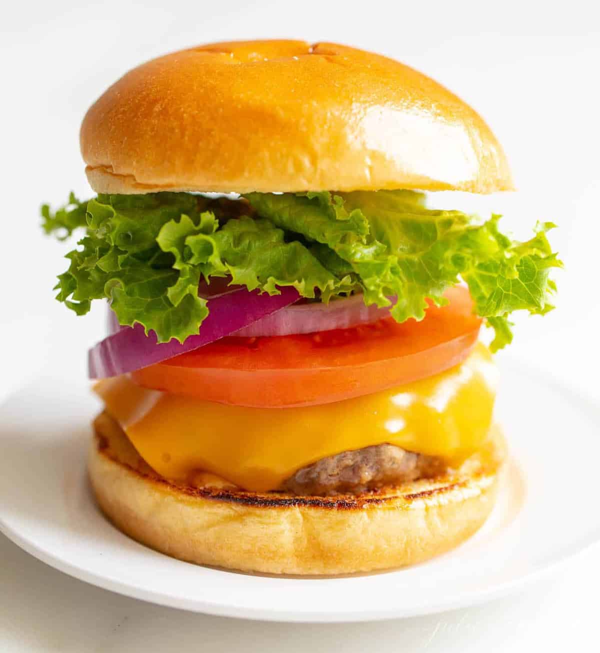 White background with a white plate featuring a classic cheeseburger, stacked with cheese, tomato, onion and lettuce.