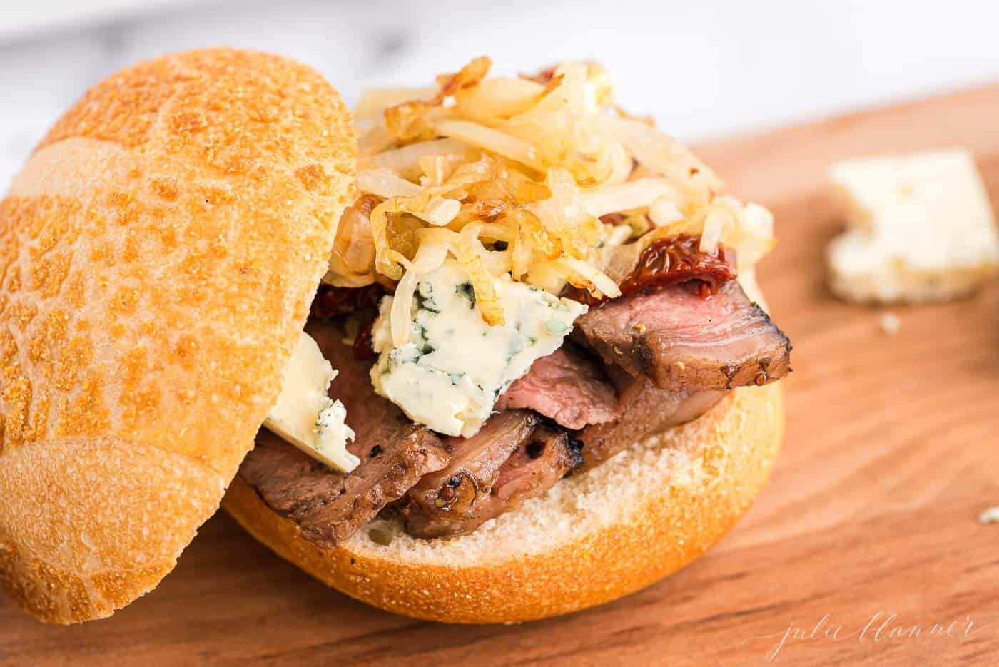 A steak sandwich with onions and blue cheese on top, bun balanced to the side.