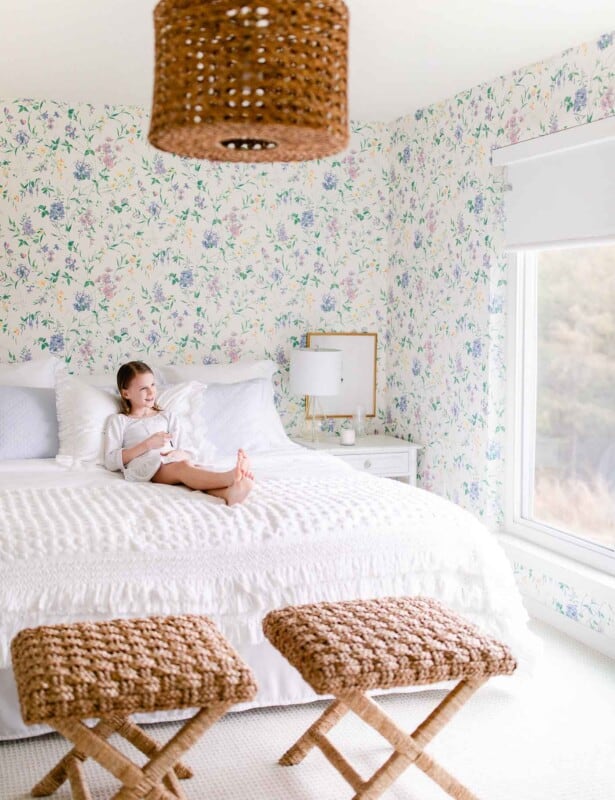 A white bedroom with floral wallpaper accents, a little girl sitting on the bed.