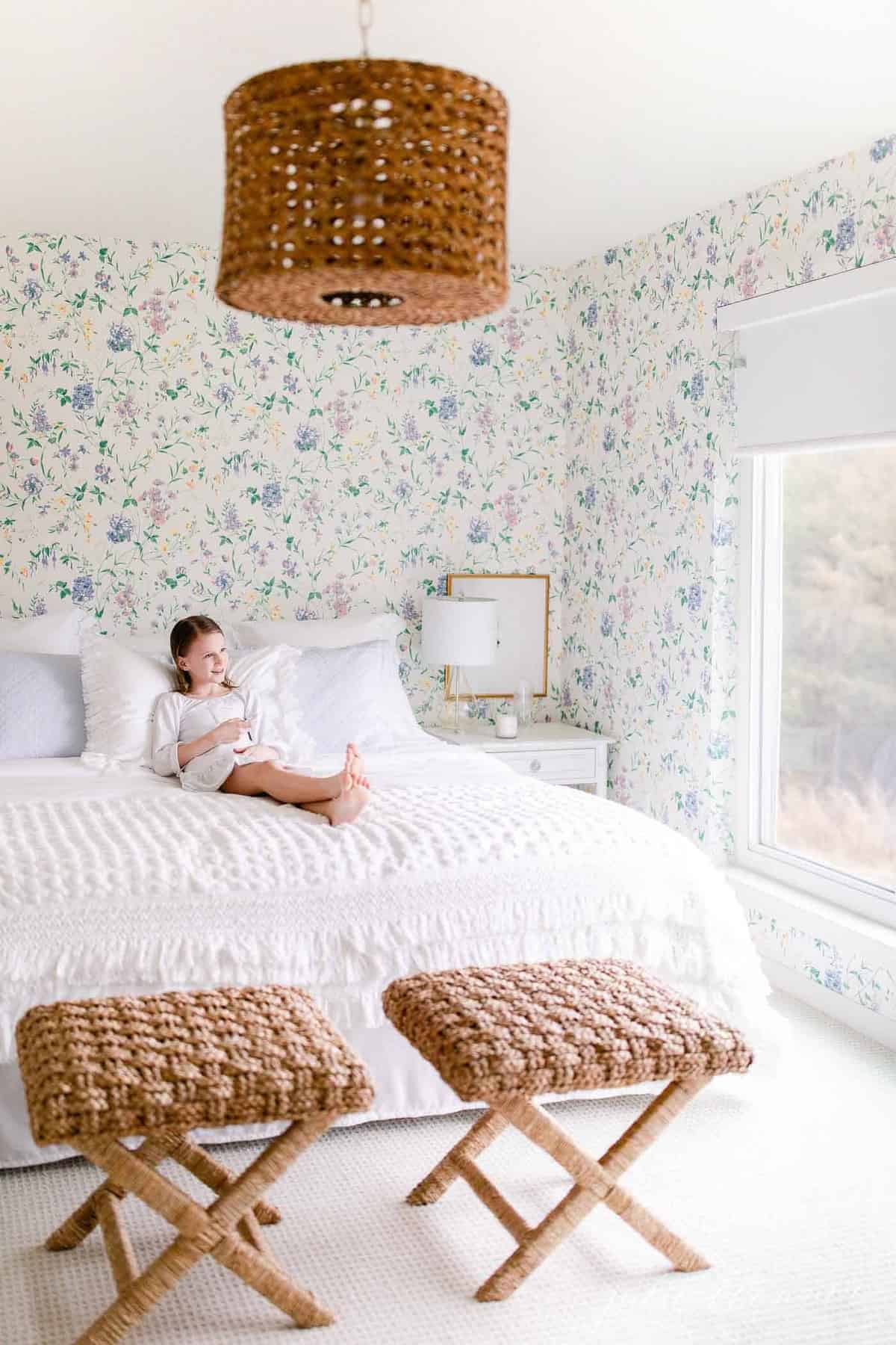 A white bedroom with floral wallpaper accents, a little girl sitting on the bed. 