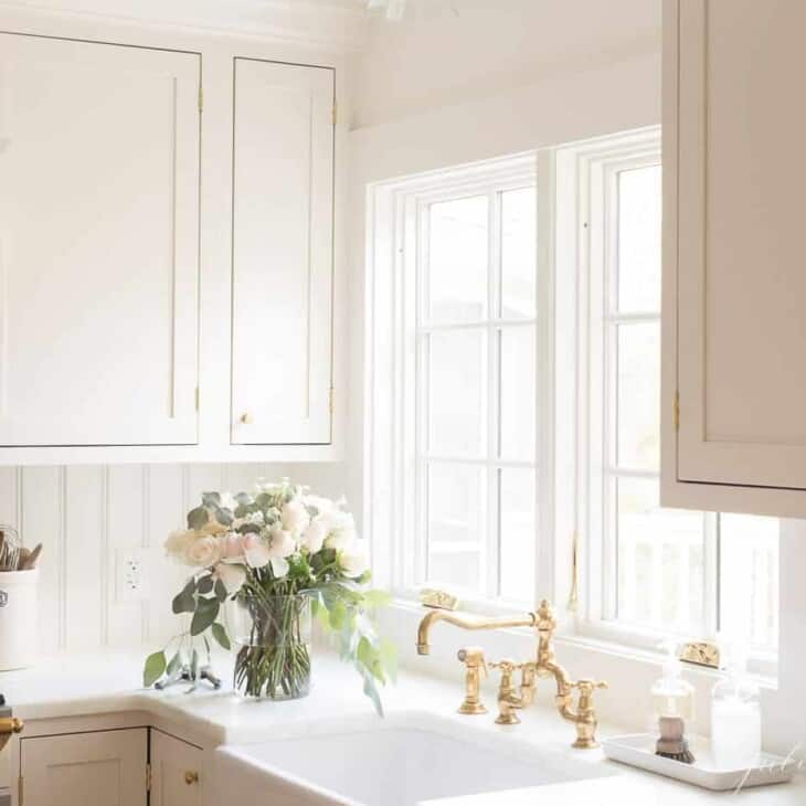 A white kitchen with a farmhouse sink, brass kitchen faucet and a vase of flowers to the side.