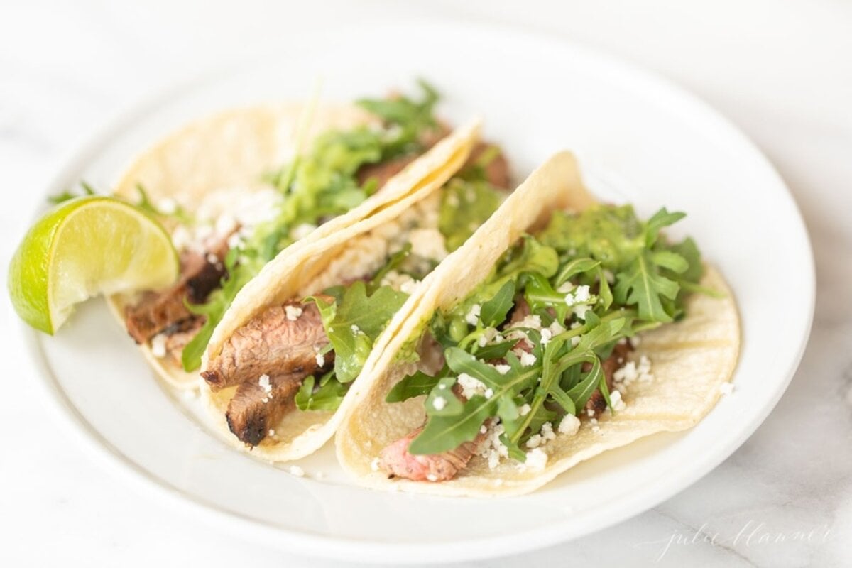 A white plate featuring three carne asada tacos topped with sauce, cheese and arugula.