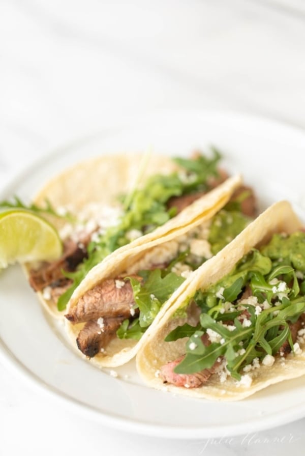 A white plate featuring three carne asada tacos topped with sauce, cheese and arugula.
