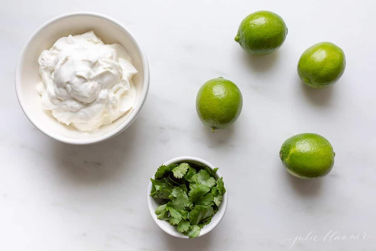 Crema de Cilantro Lime ingredients in a white bowl, with cilantro and limes to the side, on a white surface.