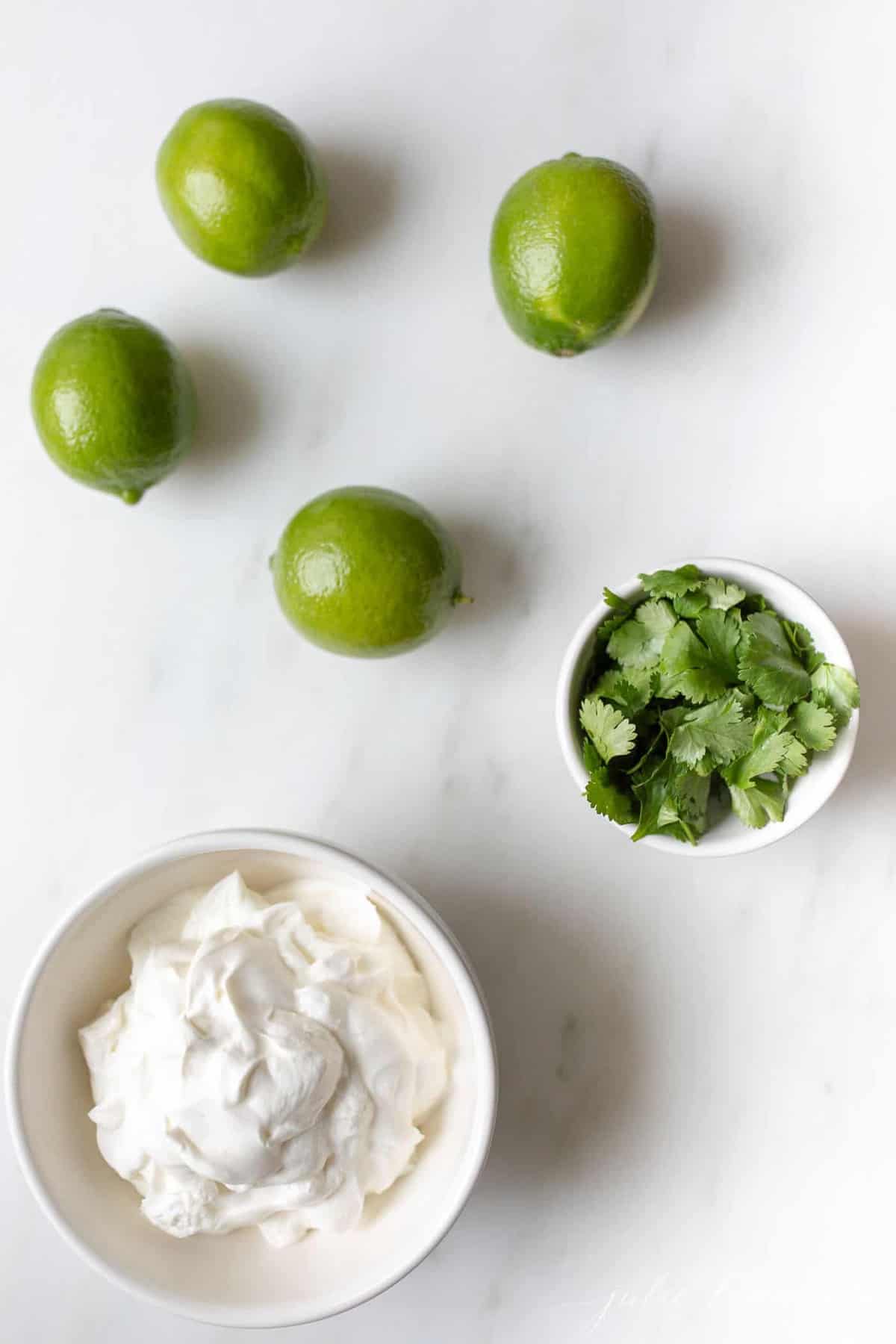 Crema de Cilantro Lime ingredients in a white bowl, with cilantro and limes to the side, on a white surface. 