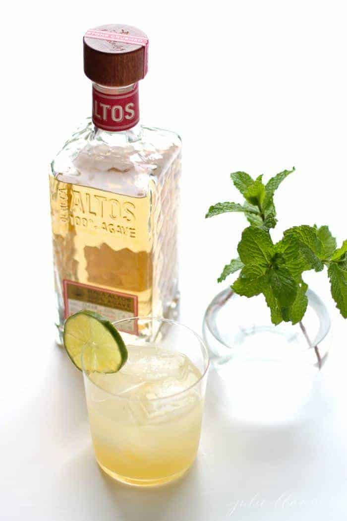 A white surface with a bottle of tequila, a sprig of mint and a margarita in a clear glass.