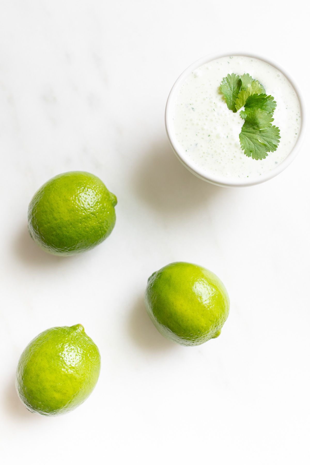 A white bowl of crema de cilantro on a white countertop, garnished with cilantro, limes to the side.