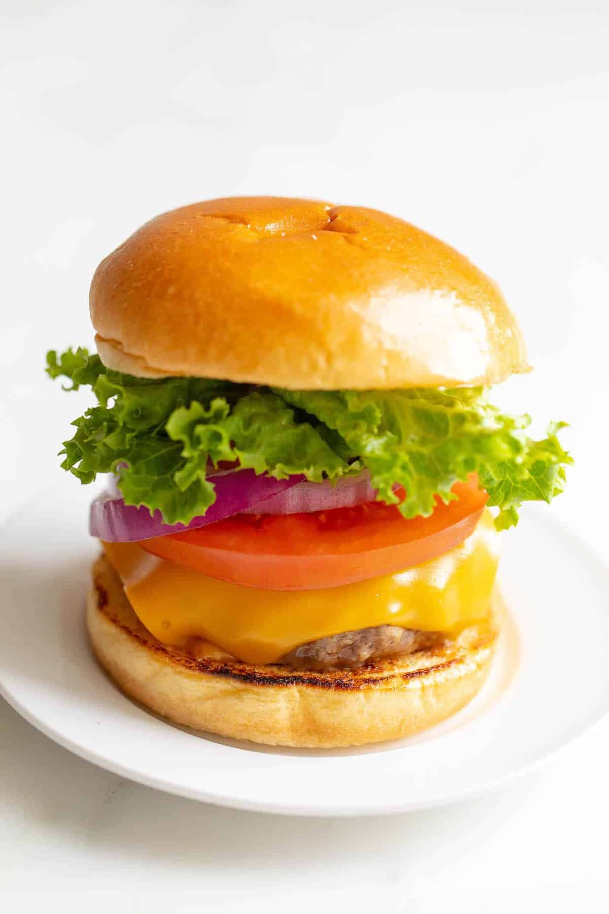 White background with a white plate featuring a classic cheeseburger, stacked with cheese, tomato, onion and lettuce.