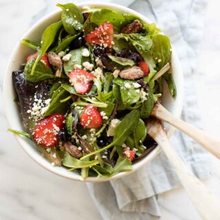 A white bowl full of a fresh pot luck salad, wooden spoons to the side..