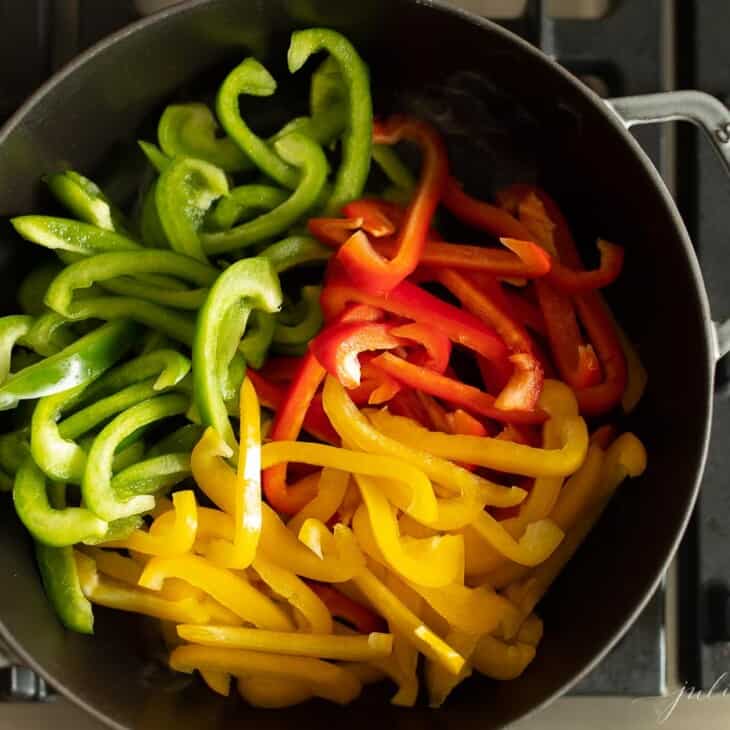 A cast iron pan on the stove, full of red green and yellow sliced peppers preparing to saute.