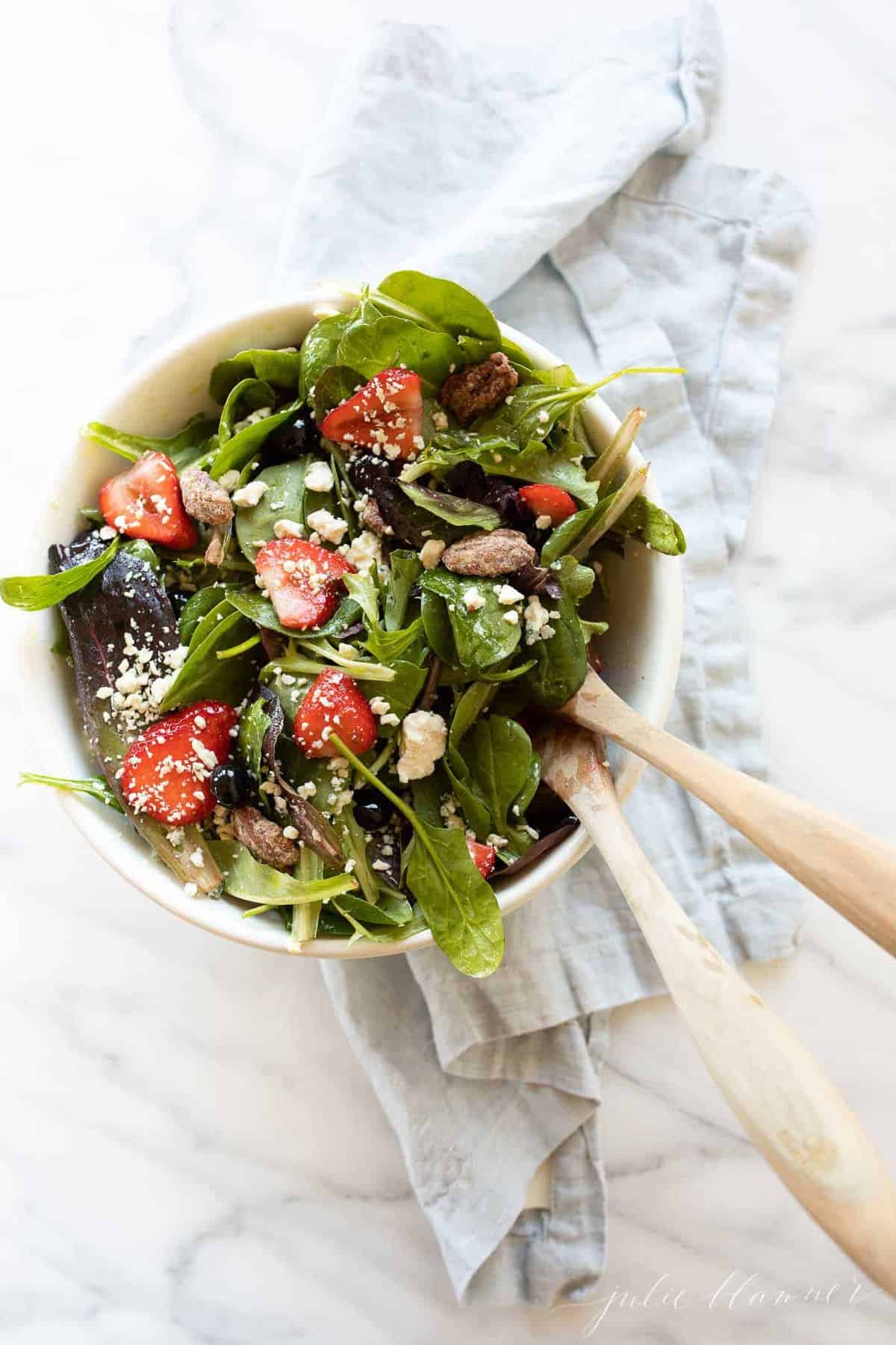 A white bowl full of a fresh pot luck salad, wooden spoons to the side..