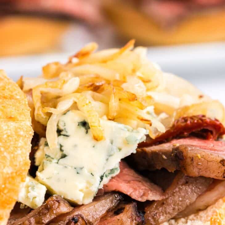 A steak sandwich with onion straws and cheese on top, top bun balanced to the side.