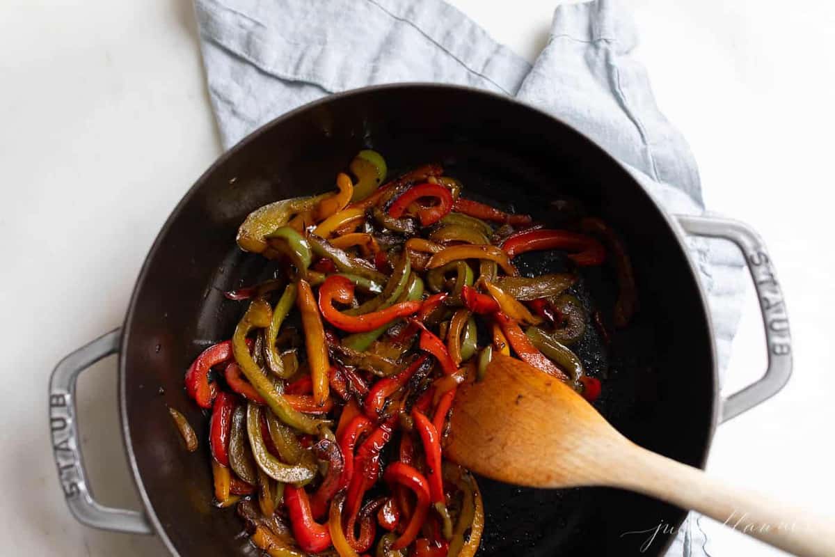 A cast iron pan full of sauteed bell peppers, wooden spoon to the side.