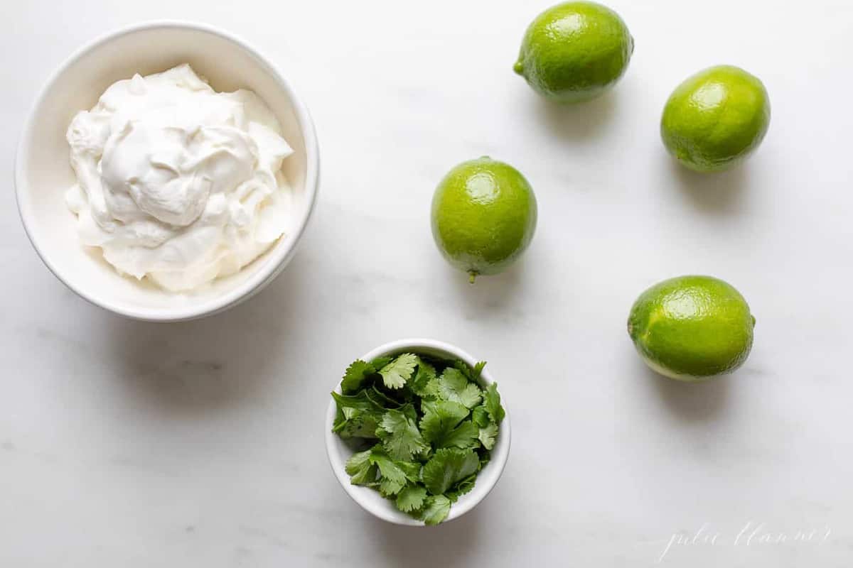 Crema de Cilantro Lime ingredients in a white bowl, with cilantro and limes to the side, on a white surface.