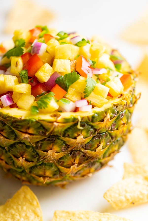 Pineapple salsa inside a pineapple bowl, on a white countertop. surrounded by tortilla chips.