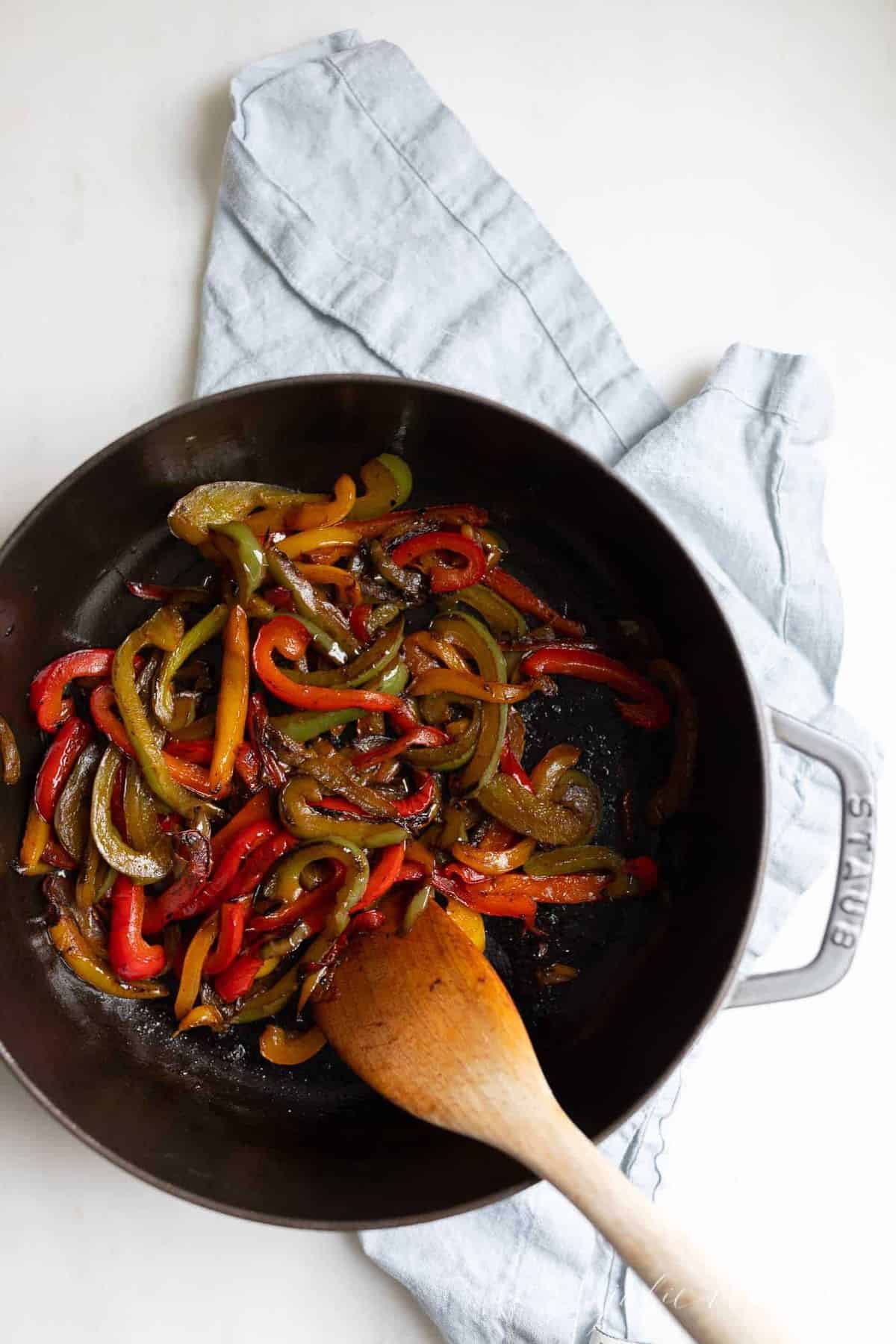 A cast iron pan full of sauteed bell peppers, wooden spoon to the side.