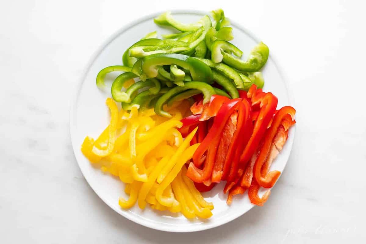 Red, green, and yellow bell peppers cut on a white plate on a white surface.