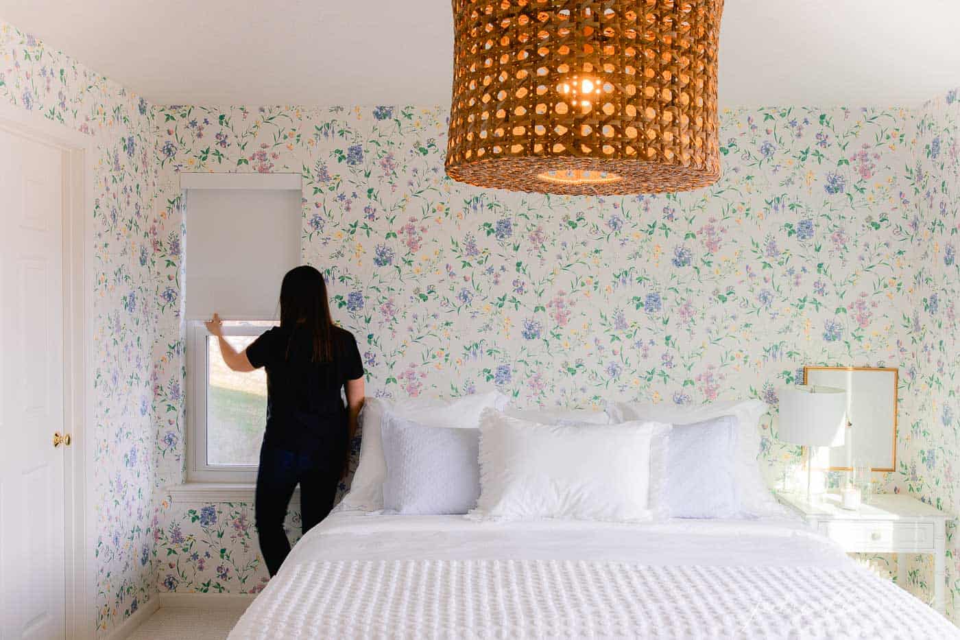 A white bedroom with wooden accents and a statement floral wallpaper, a woman pulling shades down to the side. 