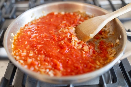A wooden spoon stirring onions in amatriciana sauce in a stainless steel skillet on a stove.