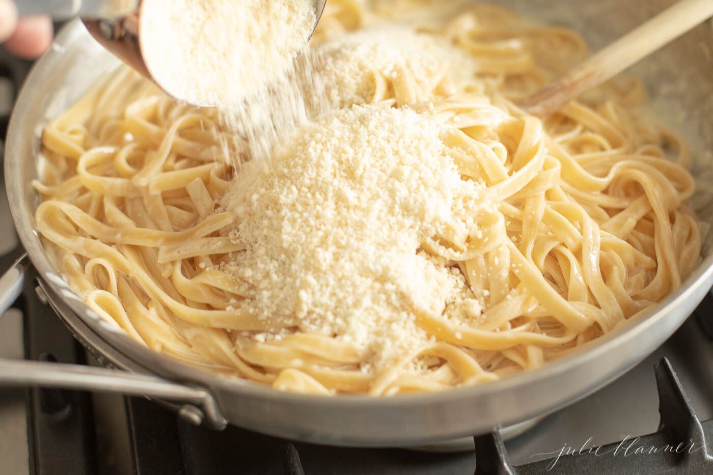 Fettuccine Alfredo in a metal pan with parmesan cheese being poured in. 