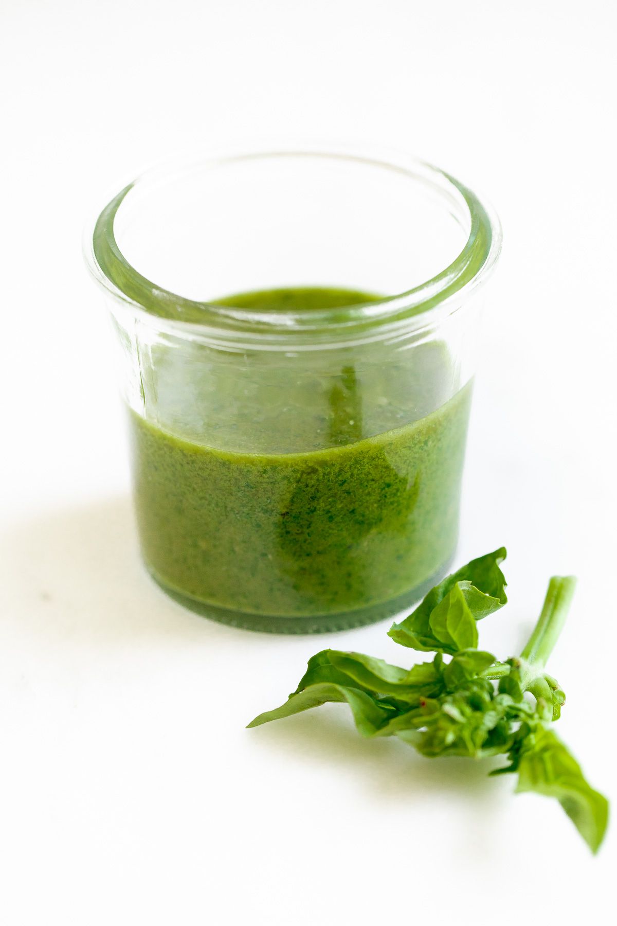 Fresh basil pesto in a small glass jar, with a stem of basil next to it on a white surface. 