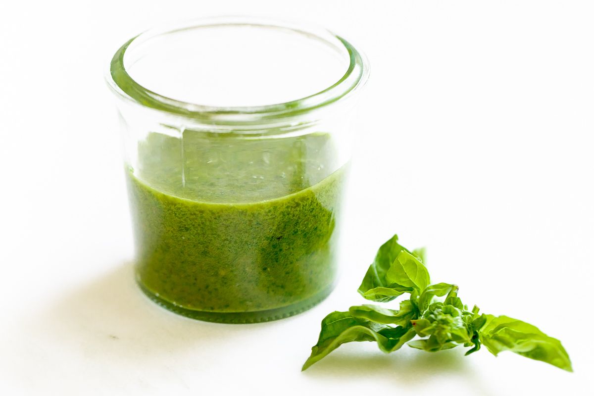 Fresh basil pesto in a small glass jar, with a stem of basil next to it on a white surface.