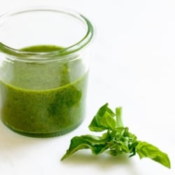 Fresh basil pesto in a small glass jar, with a stem of basil next to it on a white surface.