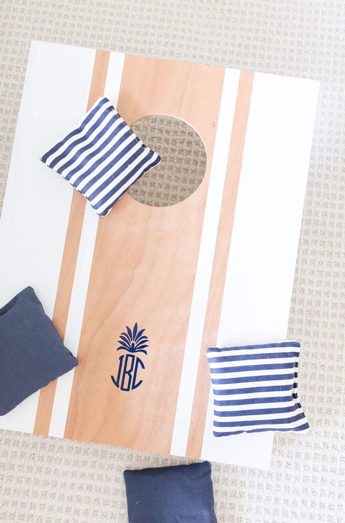 A cornhole board with a wooden surface and a white strip design with the letters "JBC" in blue at the bottom, surrounded by three striped and one plain navy blue bean bags.