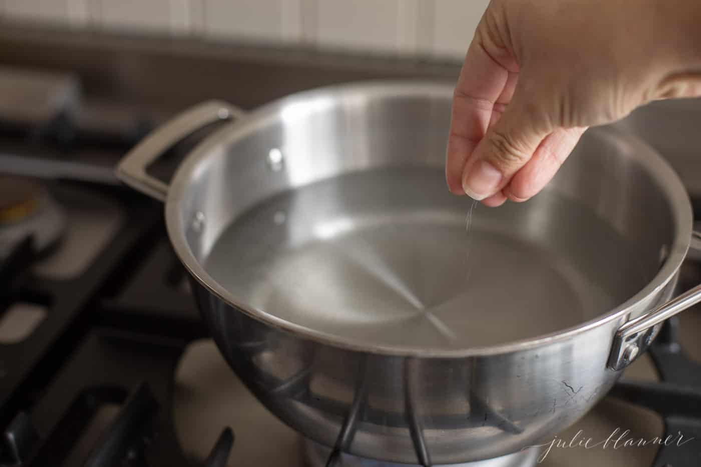 A silver pot full of water on the stove, hand reaching in with salt.
