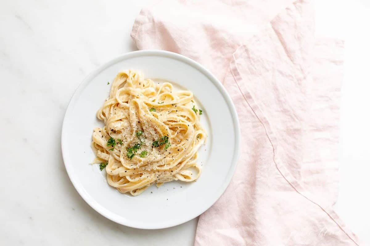 Fettuccine Alfredo on a white plate, on top of a white surface with a pink linen napkin to the side.