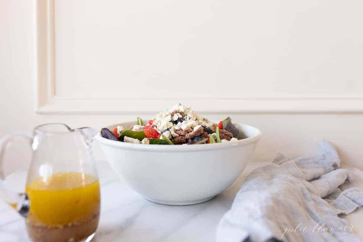 A white kitchen background with a white bowl full of salad and champagne vinaigrette in a clear pitcher. 