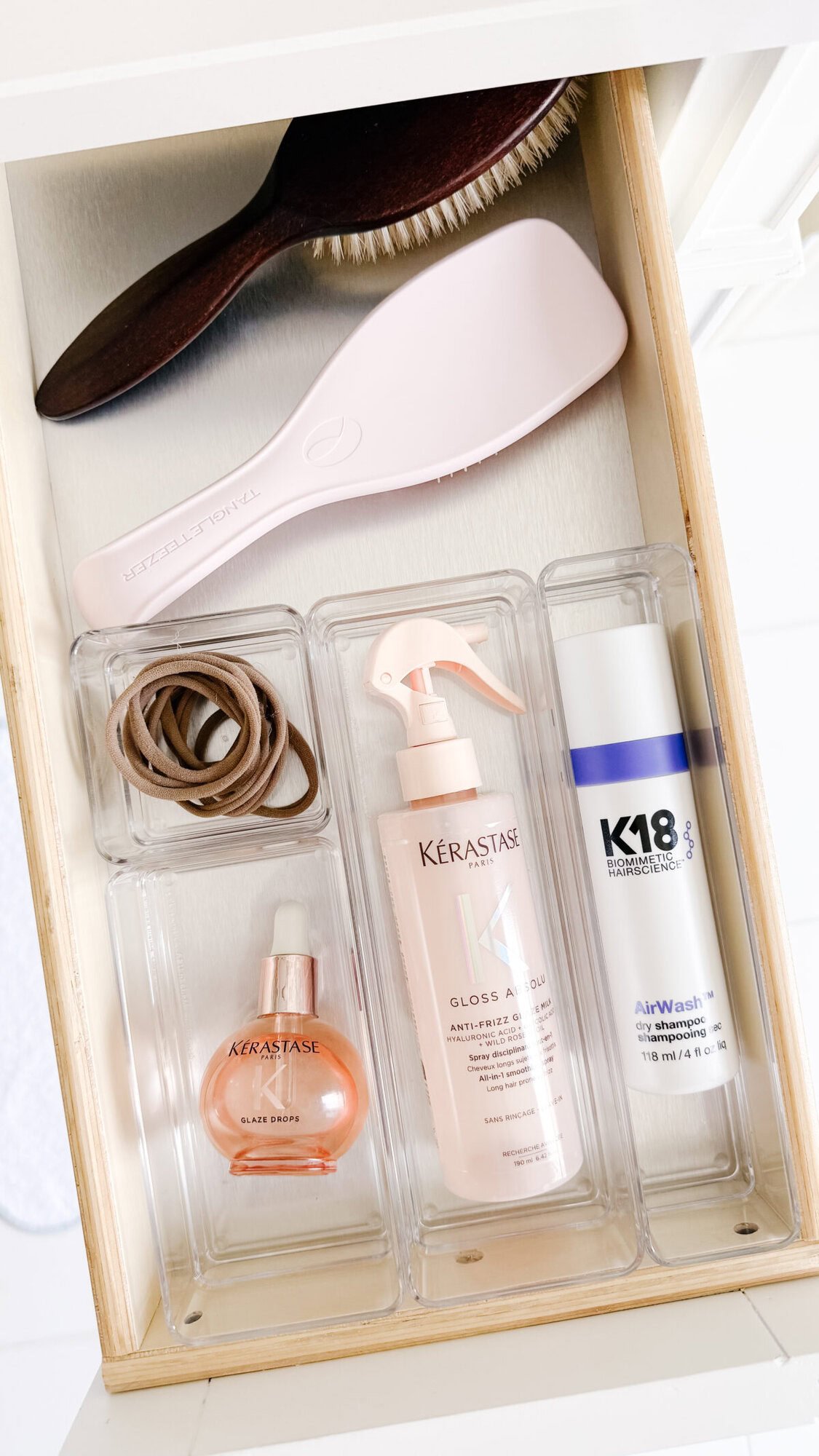 An open drawer with drawer organizers holds a hairbrush, hair tie, and three hair products—Kérastase oil, Kérastase spray, and K18 leave-in molecular repair mask—neatly arranged in clear trays.