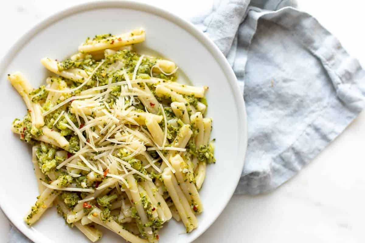 A white plate full of cheesy broccoli pasta, blue linen napkin behind.