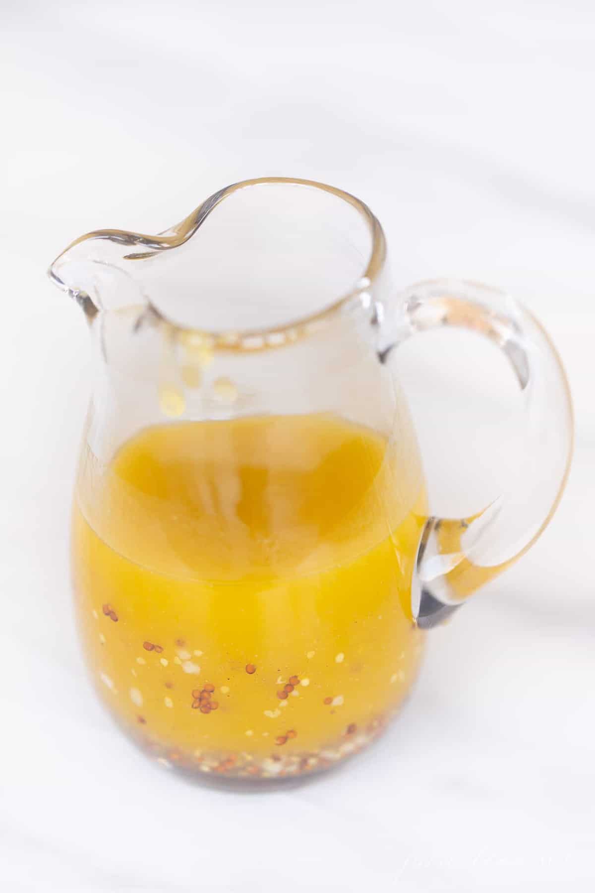 A white background with a small clear pitcher of homemade champagne vinegar dressing.